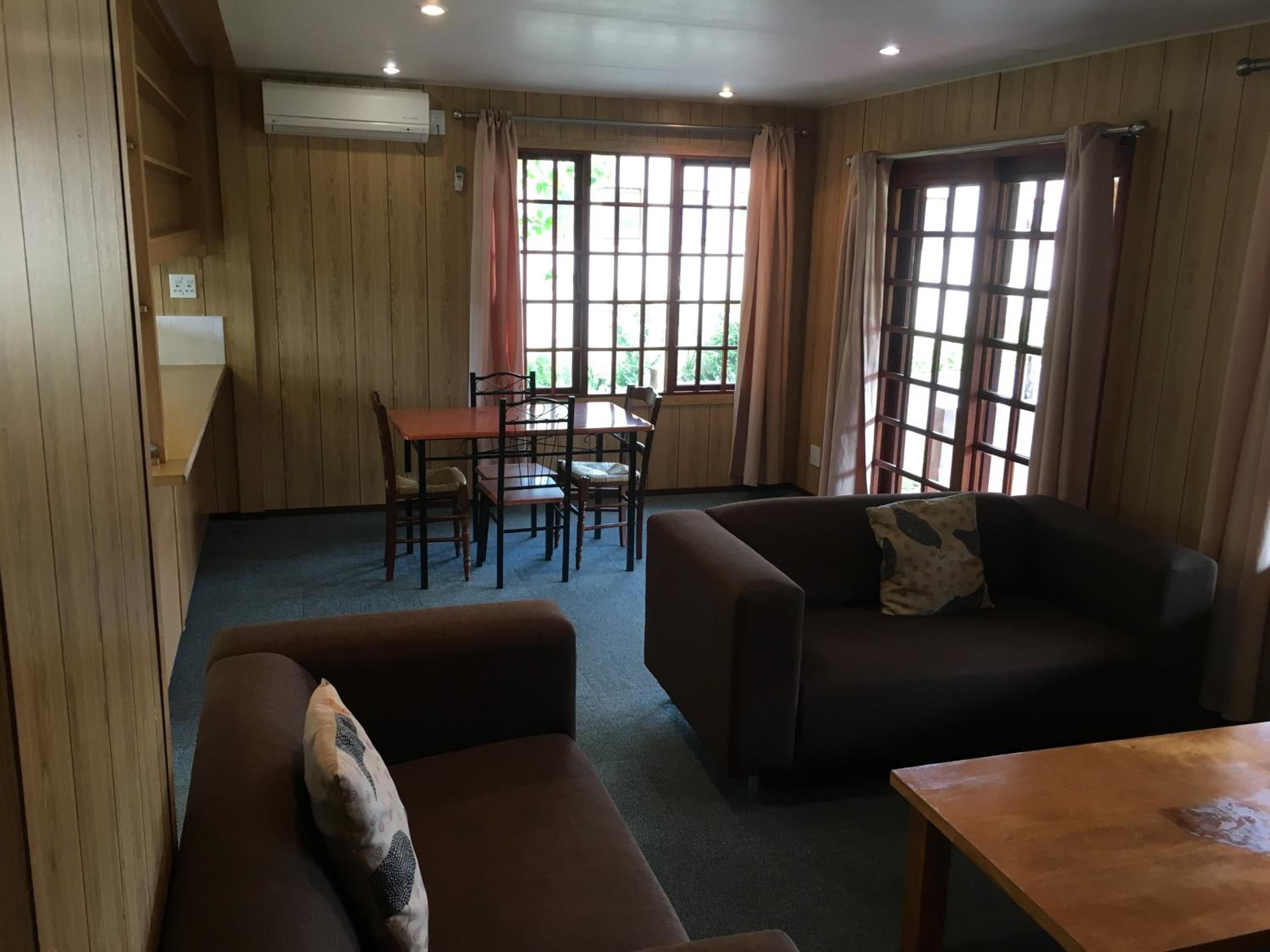 A room with a wooden paneled wall, a dining table with chairs, a sofa, and a window with curtains.