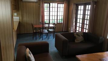 A room with a wooden paneled wall, a dining table with chairs, a sofa, and a window with curtains.