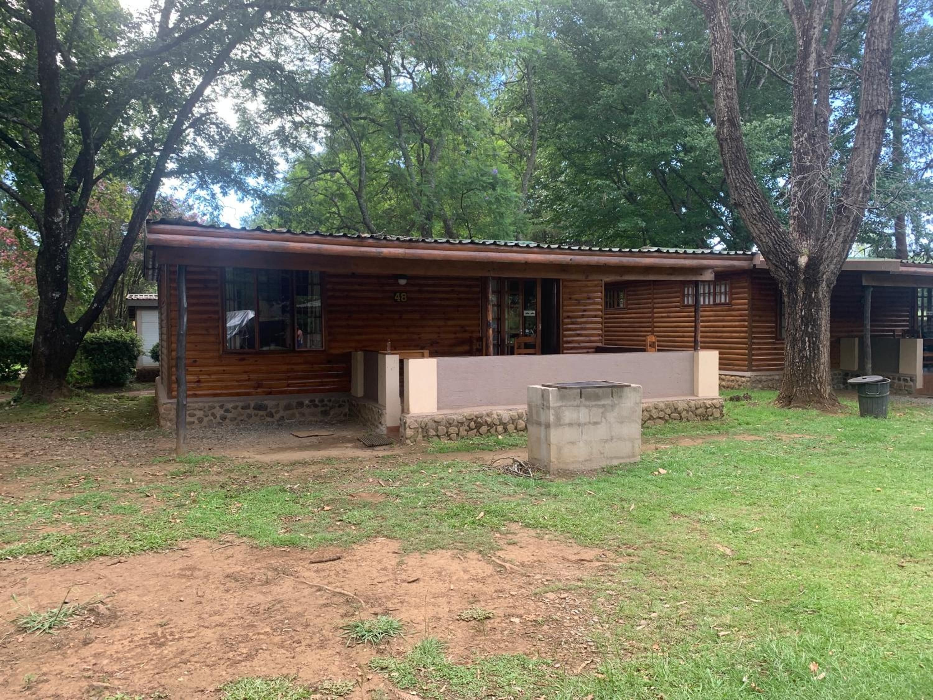 Una cabaña de madera con cimientos de piedra, una jardinera de concreto y un árbol en primer plano.