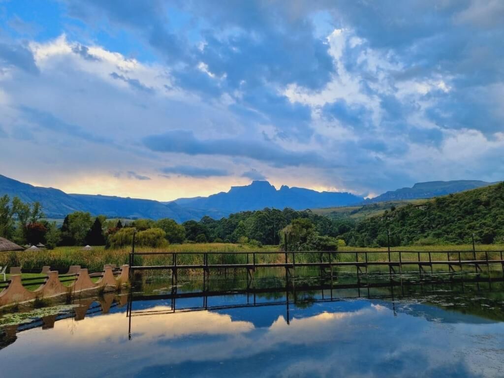 Un paysage paisible avec une étendue d’eau qui reflète le ciel, un pont en bois et des montagnes lointaines sous un ciel nuageux.