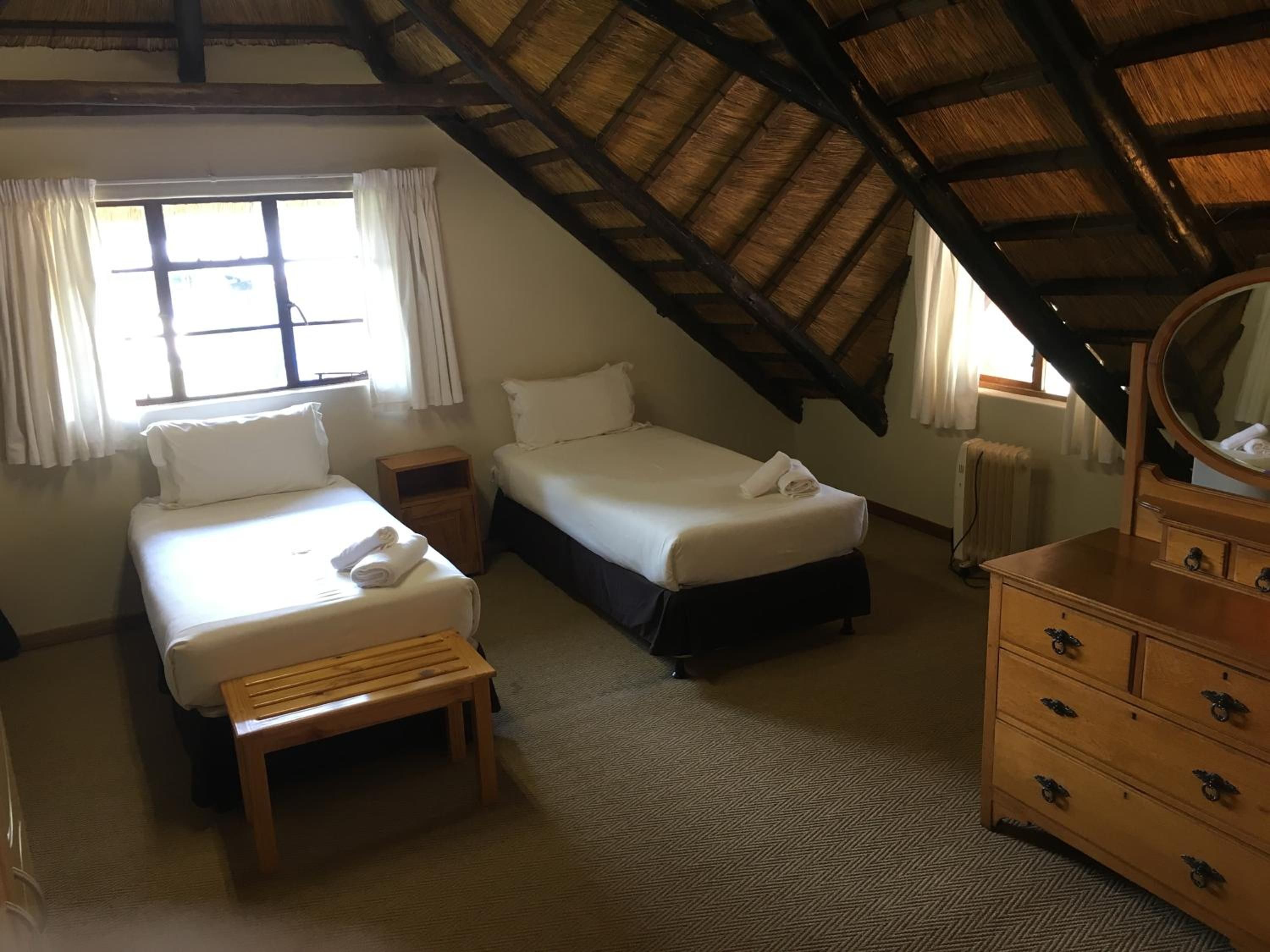 A room with two beds, a wooden bench, a dresser with drawers, and a window with curtains.
