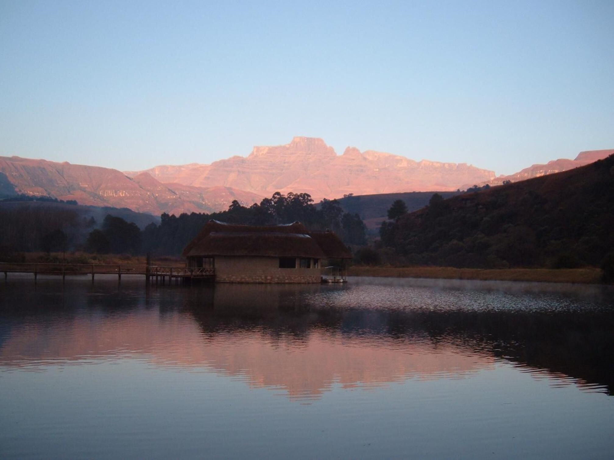 A rustic house by a calm lake with mountains in the background.