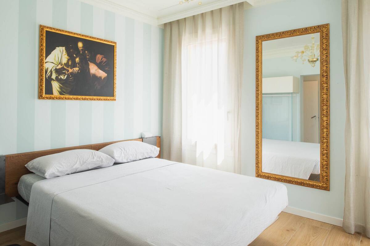 Habitación de hotel con una cama grande, un cuadro en la pared, un espejo y una ventana con cortinas.