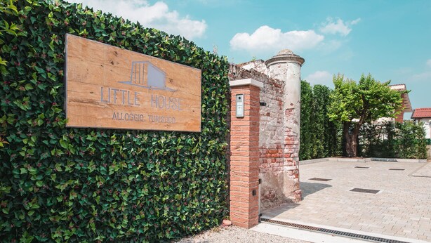 A wooden sign for 'Little House' with a brick pillar and a paved area in the background. - Little House (Campo San Martino)
