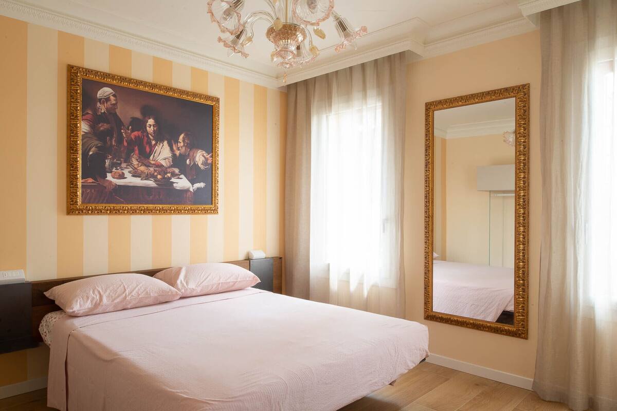 Habitación de hotel con una cama grande, un cuadro en la pared, un espejo y una ventana con cortinas.