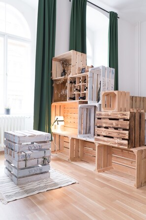 A room with wooden crates stacked on the floor, a white lamp on a wooden table, and a window with green curtains.