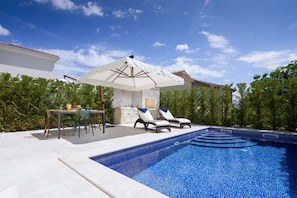 A swimming pool with lounge chairs and a table, surrounded by greenery and a clear sky.