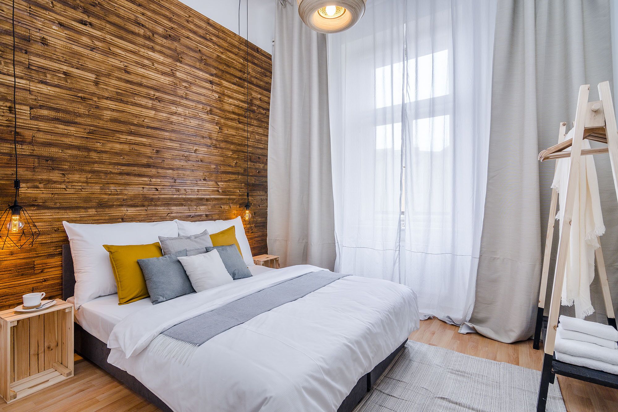 A modern bedroom with a wooden headboard, a bed with white bedding and multiple pillows, a bedside table with a cup, and a hanging light fixture.