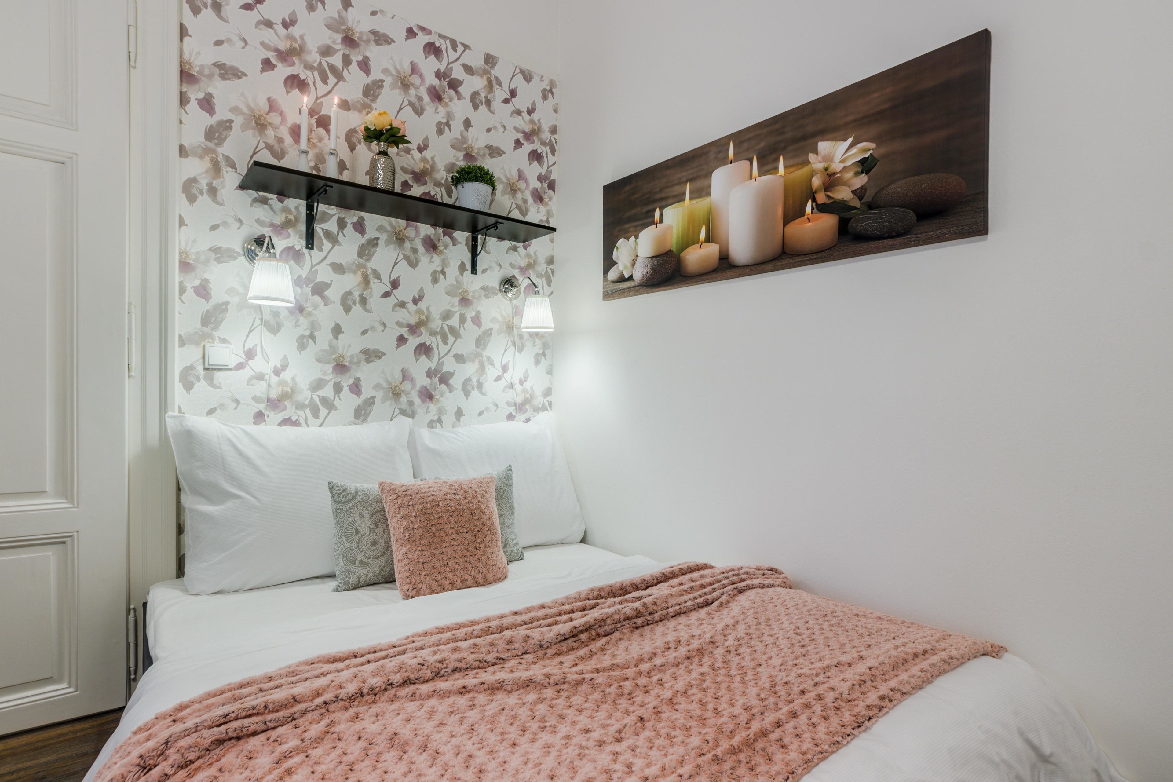 Une chambre avec un lit, une couverture rose, un papier peint à motifs floraux, une étagère avec des plantes et un tableau représentant des bougies accroché au mur.