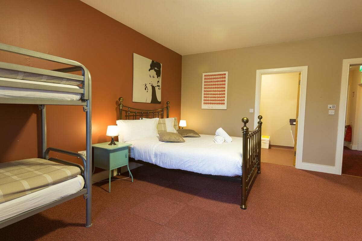 A hotel room with a bunk bed, a single bed, a desk with a lamp, and a door leading to another room.