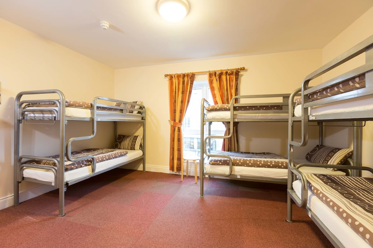 A dormitory room with bunk beds, a window with curtains, and a small table.