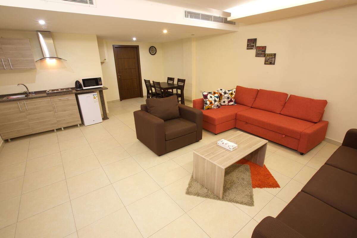A modern living room with a brown sofa, a red sectional sofa, and a coffee table. There is a kitchen area with a microwave and a refrigerator.