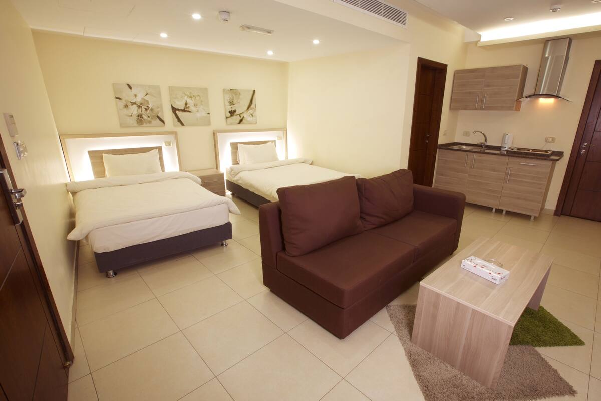 A hotel room with two beds, a brown sofa, a small wooden coffee table, and a kitchenette.