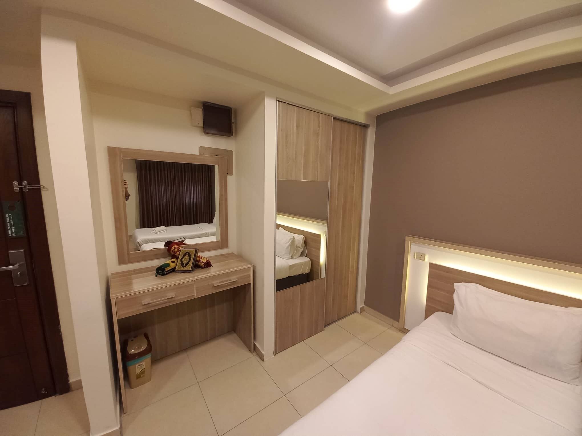 Una habitación de hotel con una cama, un escritorio con espejo, un armario y un pequeño cubo de basura.