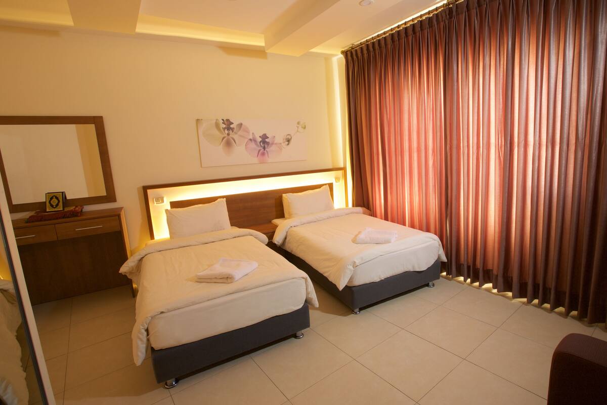 A hotel room with two beds, a wooden headboard, a nightstand, a mirror, and a large window with curtains.