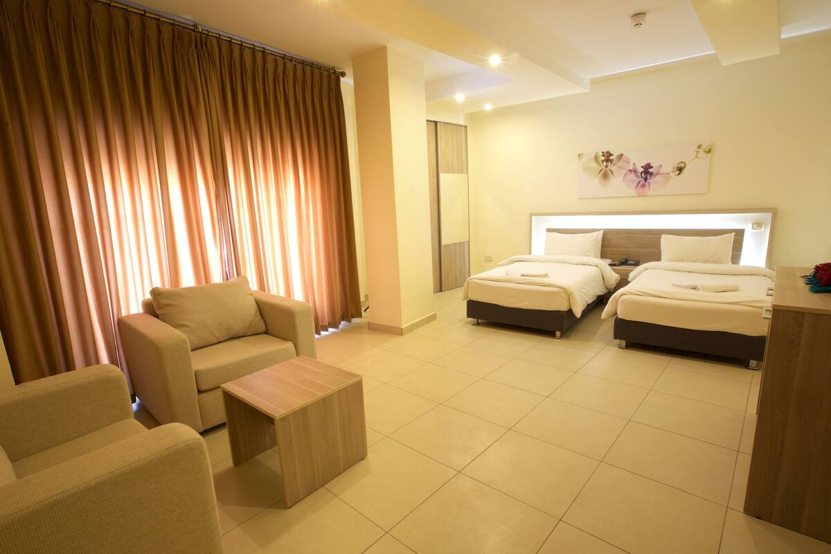 A hotel room with a sofa, two beds, a small table, and a large window with curtains.