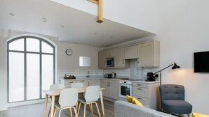 A modern kitchen with white cabinets, a dining area with a wooden table and chairs, and a grey sofa with yellow cushions. - The Gables - 2 bedroom self catered cottage apartment (BARNETBY)