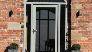 A brick building with a white-framed door and a small arched window above it. There are two potted plants on either side of the door. - The Gables - 2 bedroom self catered cottage apartment (BARNETBY)