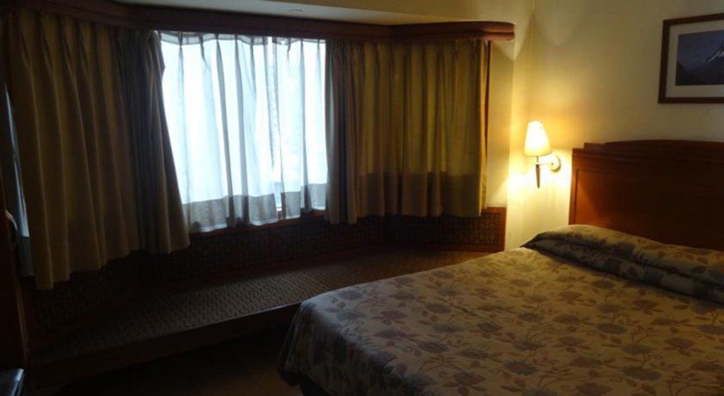 A hotel room with a bed, a window with curtains, a nightstand with a lamp, and a headboard.