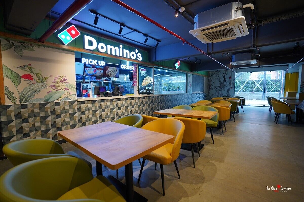 En Domino’s-restaurang med en inredning som inkluderar en disk för avhämtningsbeställningar, gula och gröna stolar samt en vägg med ett mönster.