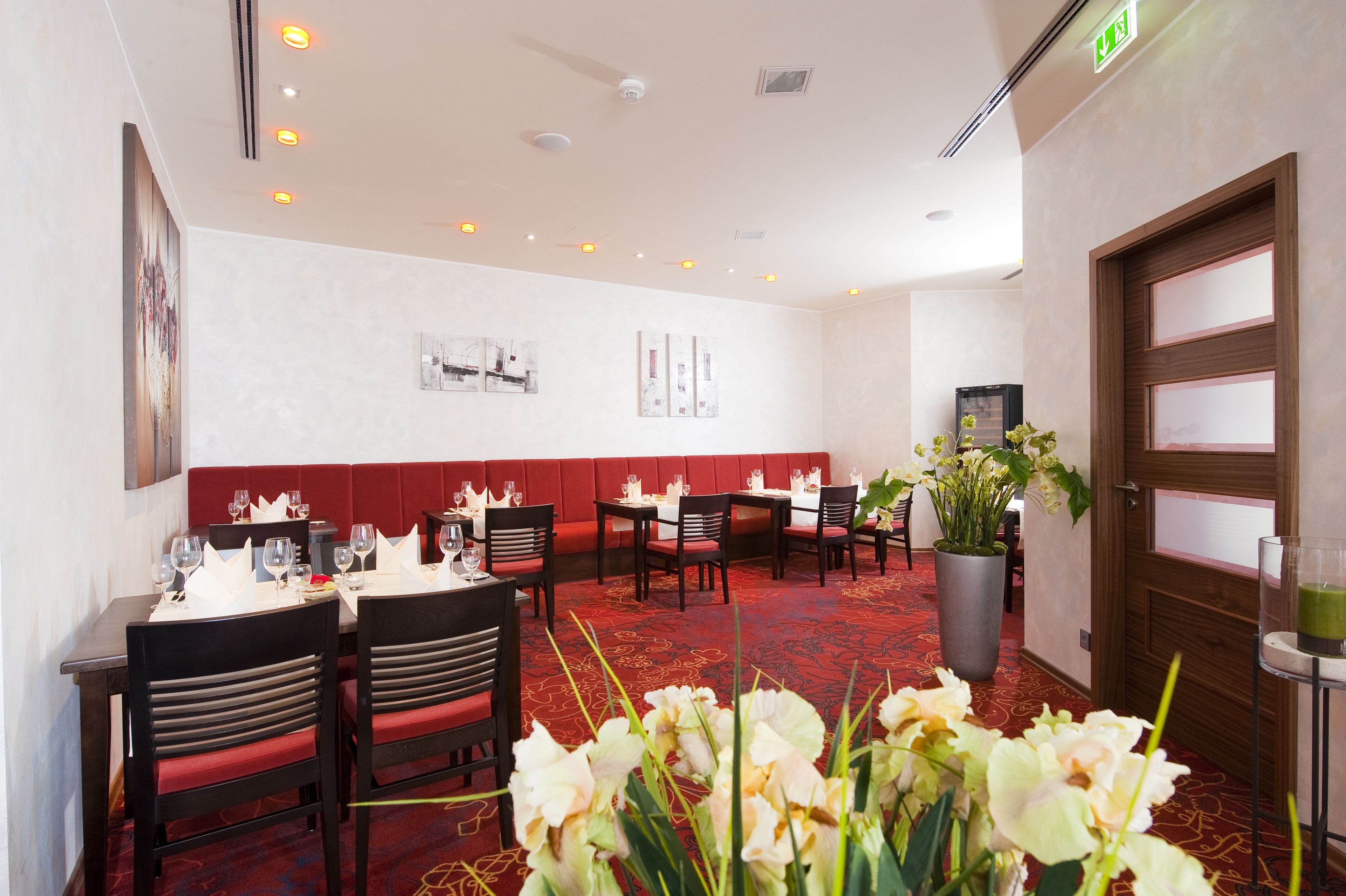 A restaurant with red cushioned booths, wooden tables, and chairs. There are potted plants and a large vase of flowers on the tables.