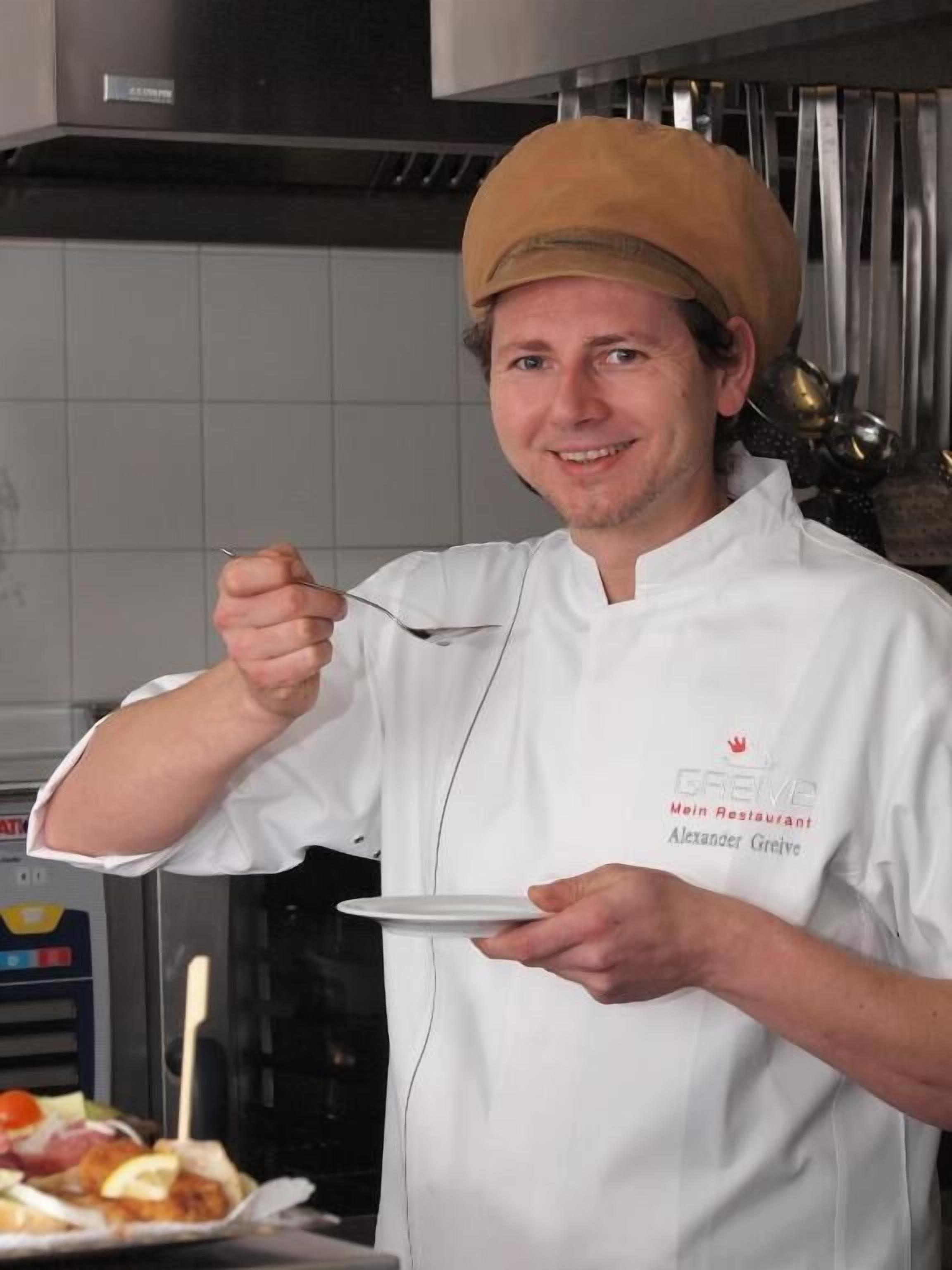 A chef in a white uniform with a name tag, holding a plate and a spoon, in a kitchen setting with stainless steel appliances and a tiled backsplash.
