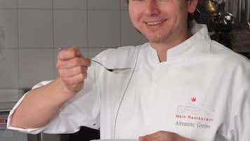 A chef in a white uniform with a name tag, holding a plate and a spoon, in a kitchen setting with stainless steel appliances and a tiled backsplash.