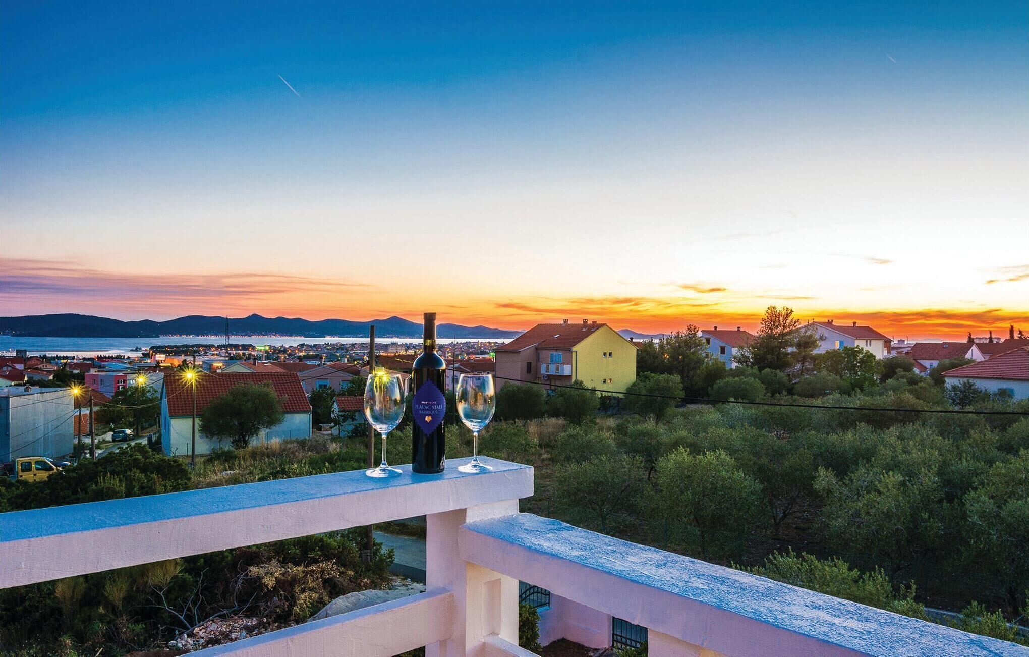 A balcony with two wine glasses and a bottle, overlooking a coastal town at sunset.