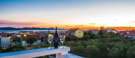 A balcony with two wine glasses and a bottle, overlooking a coastal town at sunset.