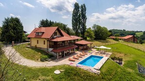 Une maison à deux étages avec un toit recouvert de tuiles rouges, une piscine et un patio aménagé avec des chaises longues.
