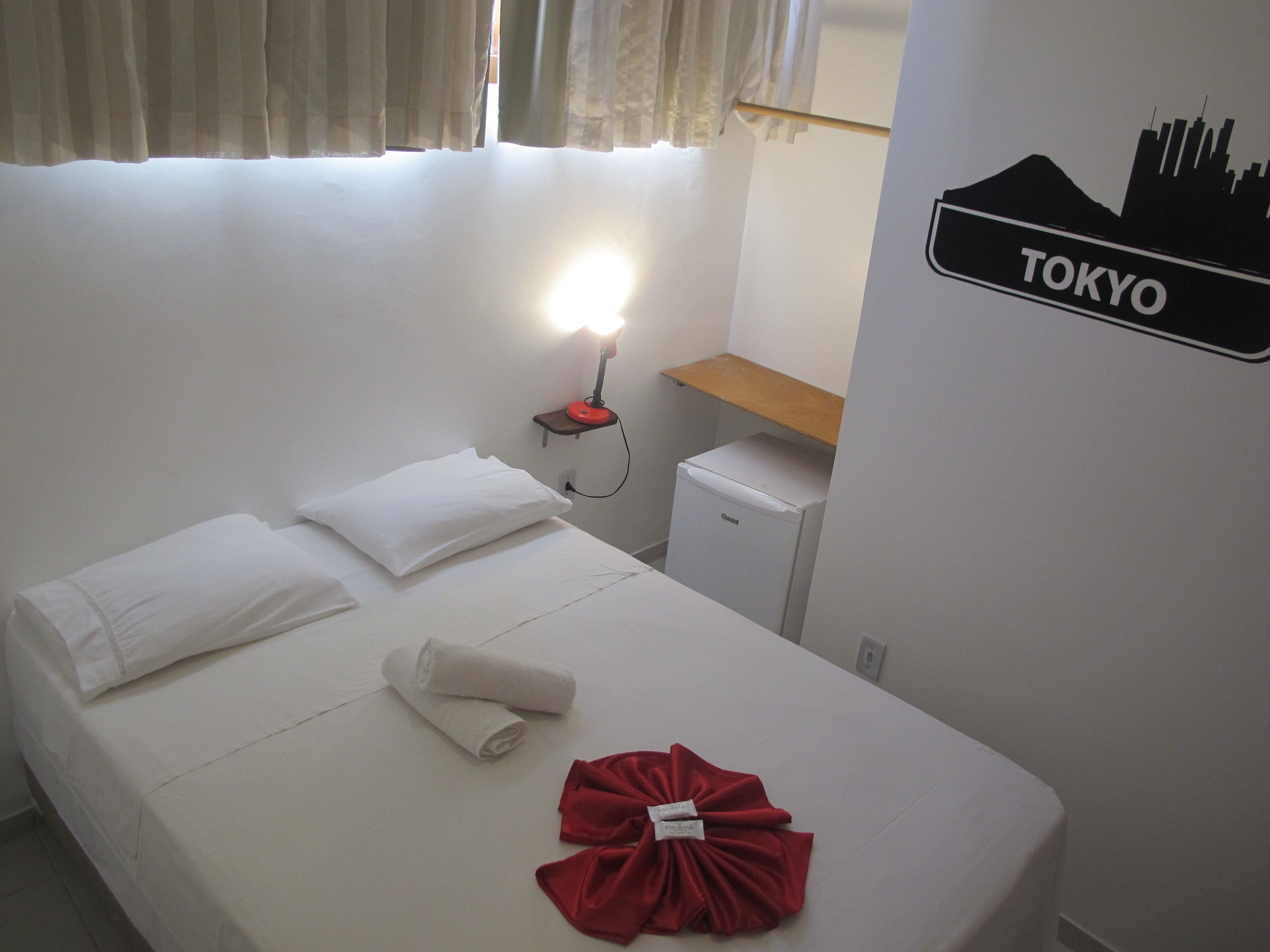 Hotelkamer met een bed, nachtkastje met lamp, kleine koelkast en een bordje dat aangeeft dat de locatie Tokio is.