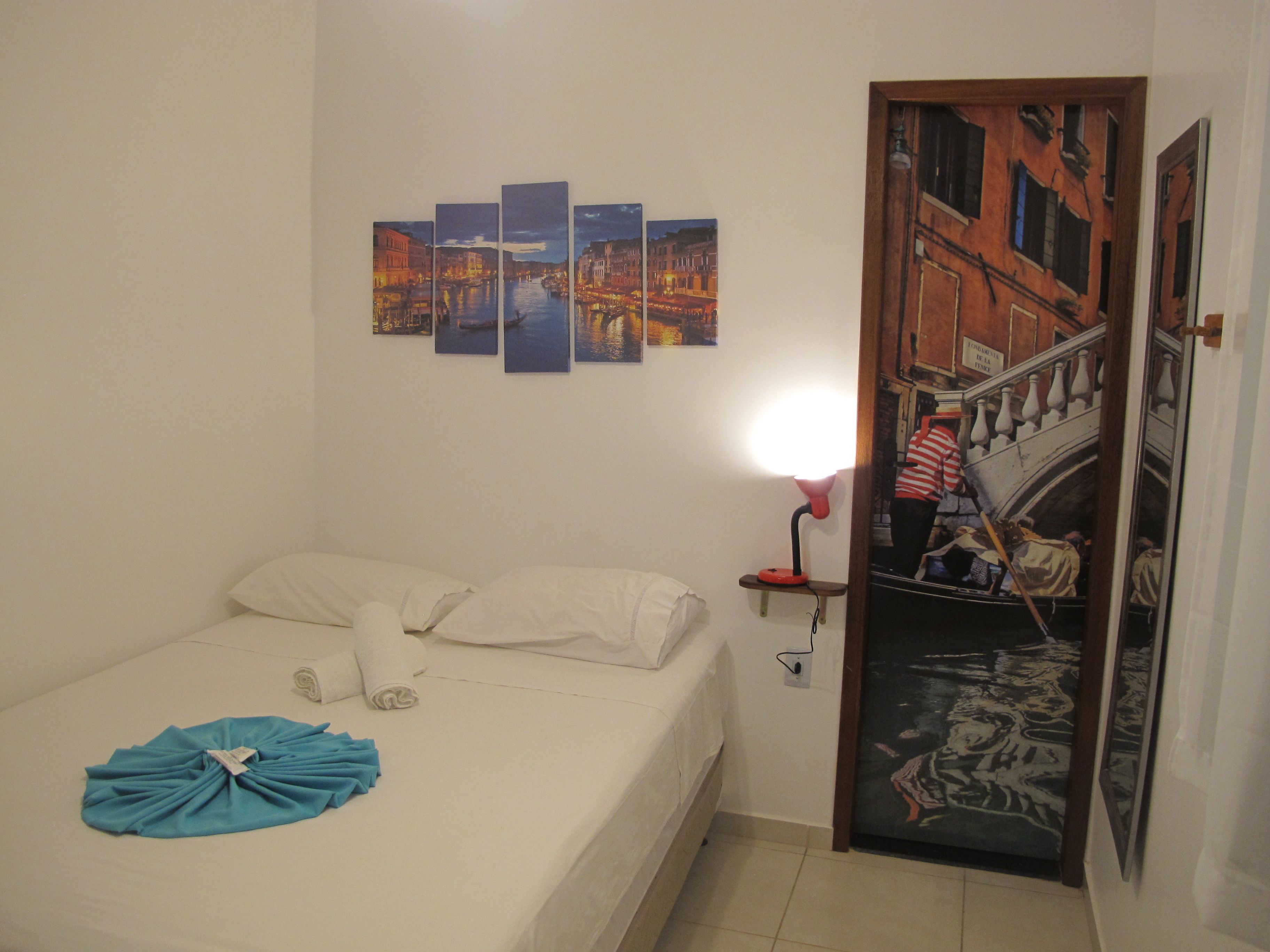 A hotel room with a bed, a wall-mounted artwork depicting a cityscape, a night lamp, and a door with a mural.