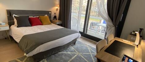 A hotel room with a bed, a desk with a laptop, a window with curtains, and a view of the outside.