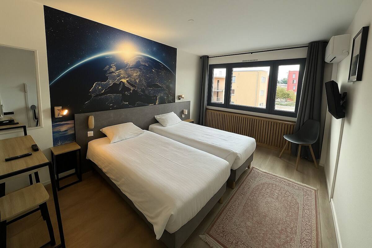Une chambre d’hôtel avec deux lits, un bureau, une chaise, une grande fenêtre donnant sur des immeubles et un mur orné d’une œuvre d’art sur le thème de l’espace.