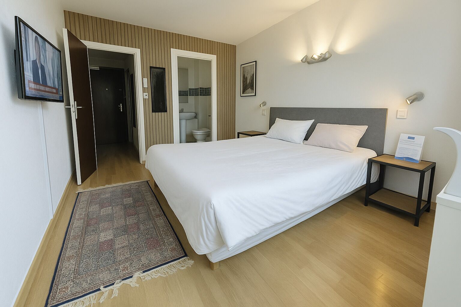 A hotel room with a large bed, a bedside table, a nightstand, a wall-mounted TV, and a door leading to a bathroom.