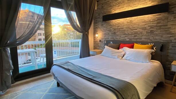 A modern bedroom with a large bed, a view of the outdoors, and a stone wall. - Savoie Hotel (Saint-Julien-en-Genevois)