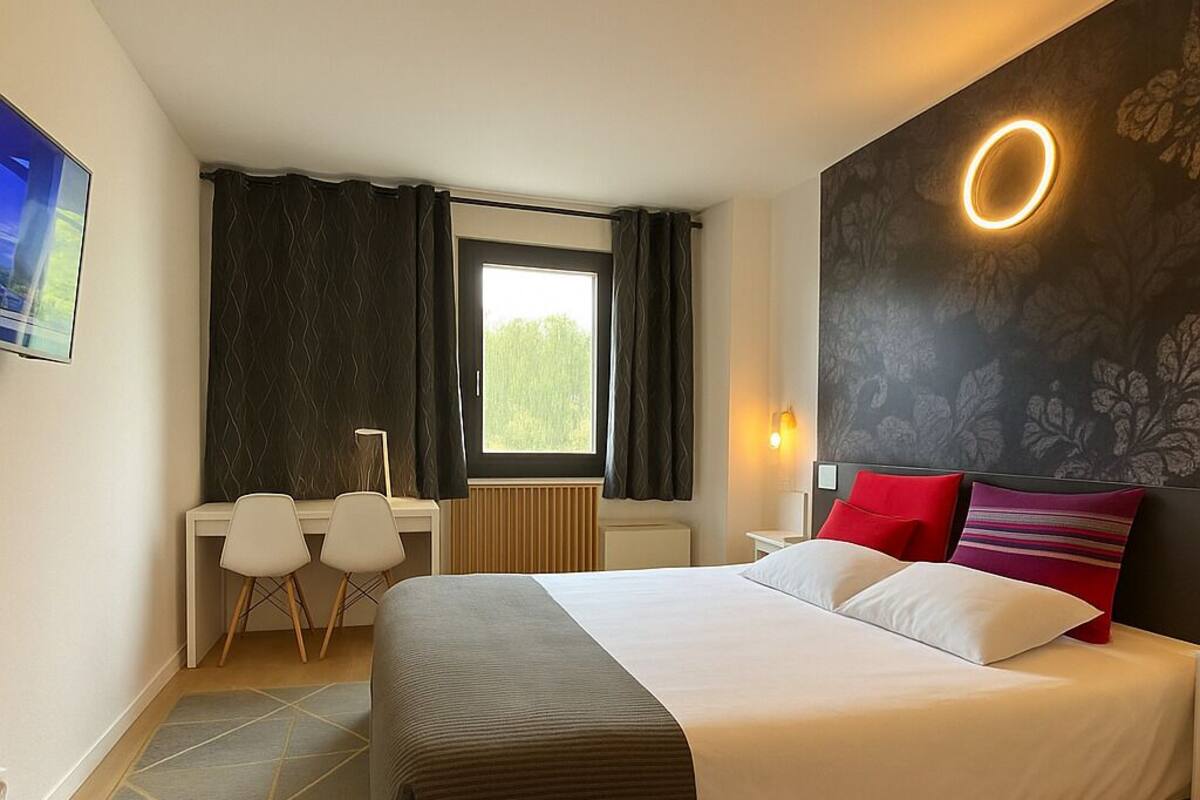 Une chambre d’hôtel comprenant un lit, un bureau, deux chaises, une fenêtre avec des rideaux et un luminaire fixé au mur.