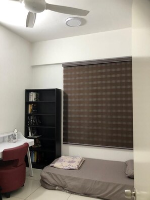 Une petite chambre à coucher, de taille réduite, comprenant un lit simple, un bureau avec une chaise, une bibliothèque et une fenêtre avec des stores.