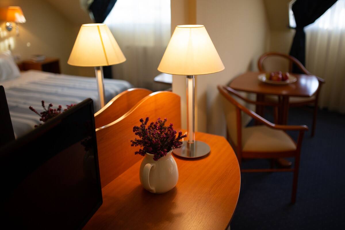 Habitació d'hotel amb llit, dues làmpades de nit, una cadira de fusta, taula petita amb un plat de fruita i un vas amb flors sobre una taula auxiliar de fusta.