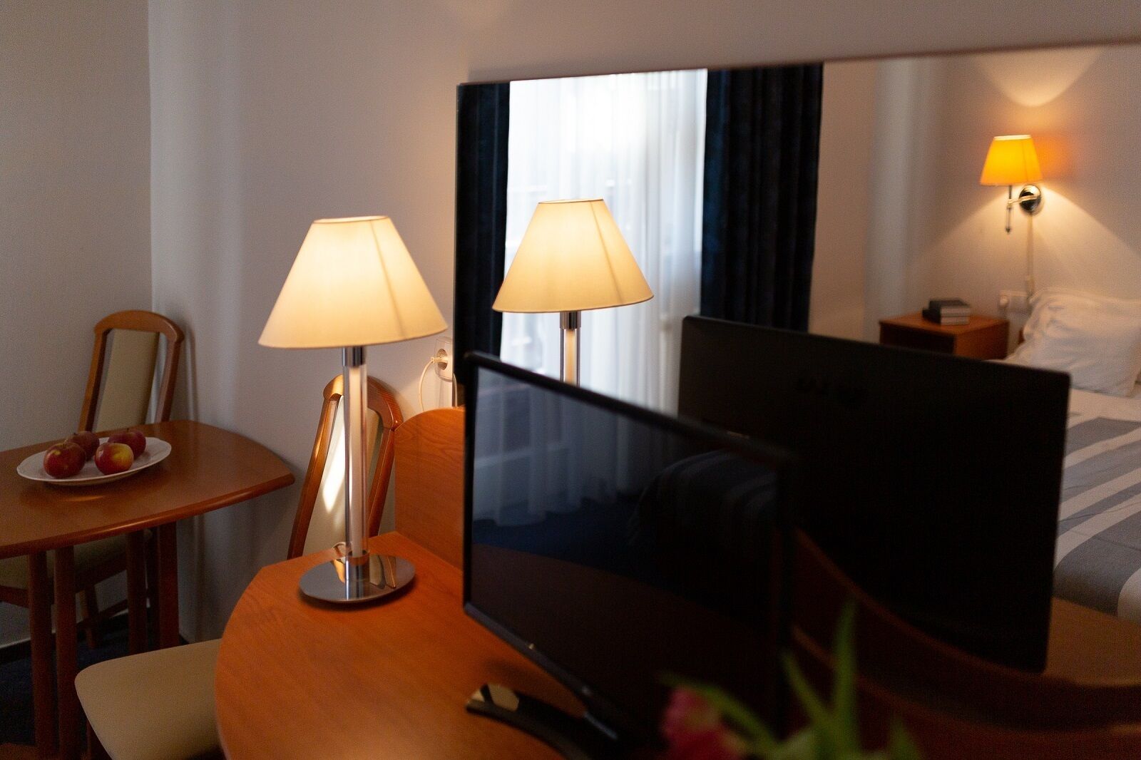 Habitación de hotel con una cama, una mesita de noche con lámpara, un televisor sobre un mueble y una mesa de comedor con manzanas.