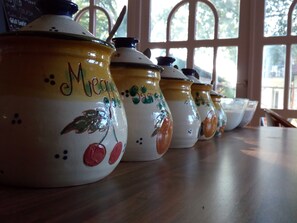 Une rangée de récipients en céramique ornés de motifs de fruits, posés sur une table en bois près d’une fenêtre.