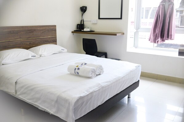A neatly made bed with white linens and rolled towels, a wooden headboard, a small desk with a lamp, and a window with a view of the outside.