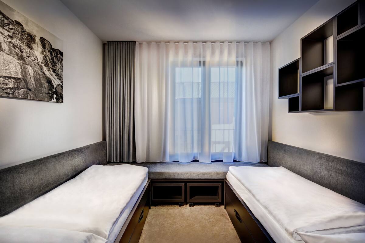 Ein Hotelzimmer mit zwei Einzelbetten, einem großen Fenster mit transparenten Vorhängen, einem Gemälde an der Wand und eingebauten Regalen.