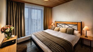Habitación de hotel con techo de madera, una cama grande con ropa de cama a rayas, una mesita de noche con una lámpara, un jarrón con flores y vistas de la ciudad a través de cortinas traslúcidas.