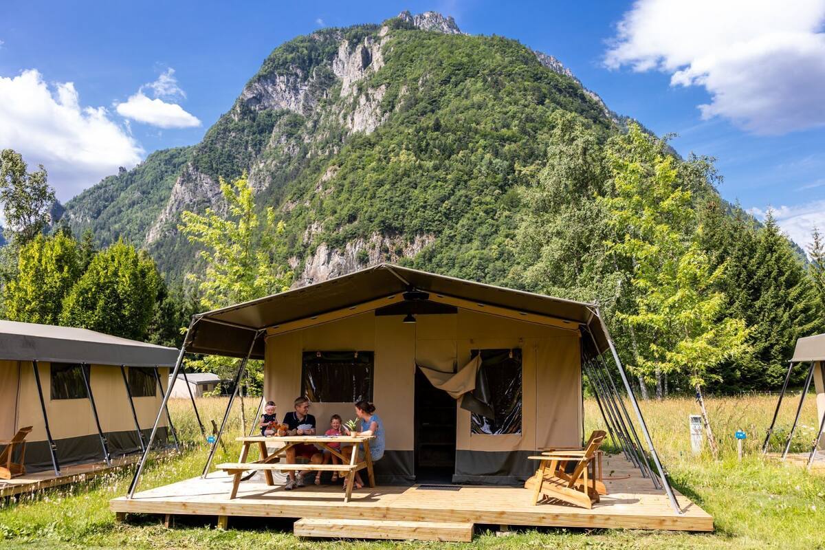 A camping site with a large tent and picnic table in front, surrounded by lush greenery and a mountain in the background.