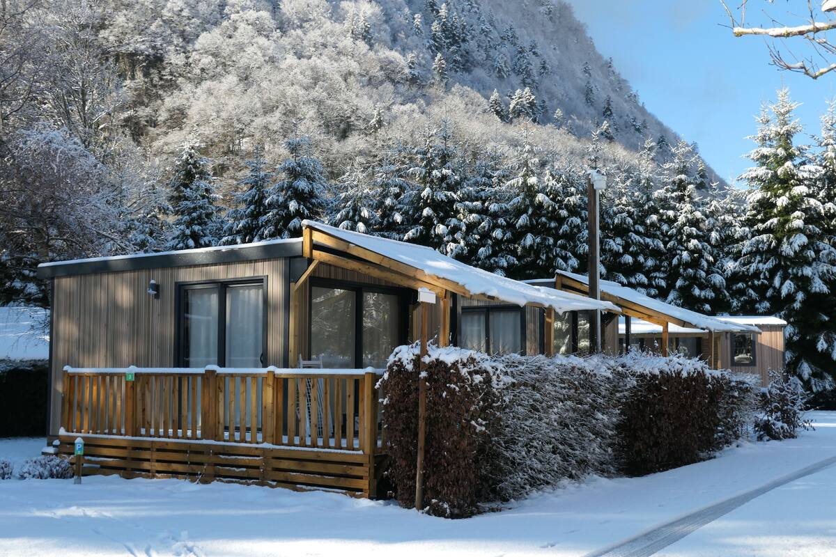 A wooden cabin with a balcony, surrounded by snow-covered trees and a mountain in the background.