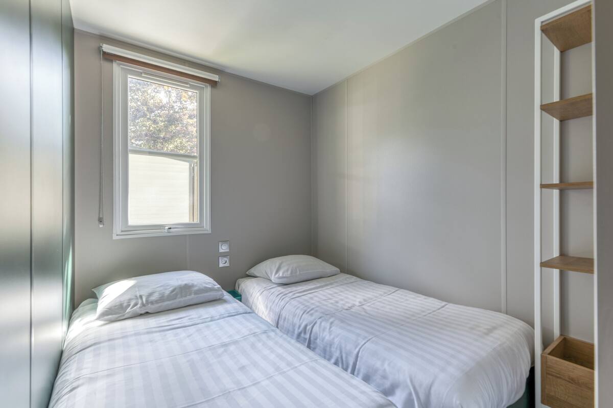 A small, single-room with two beds, a window, and a built-in shelf.