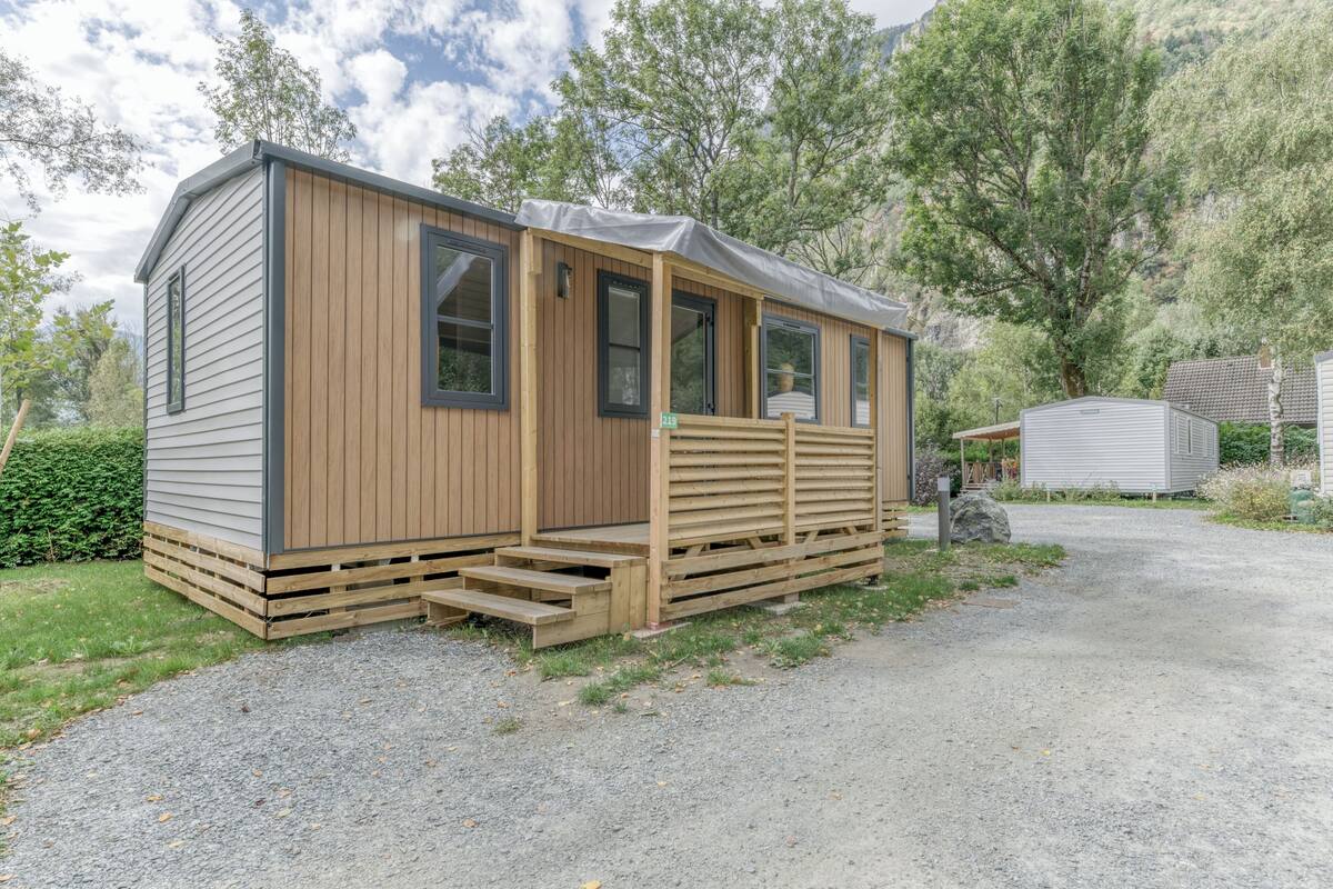 A small wooden cabin with a porch, a gravel driveway, and a forested backdrop.