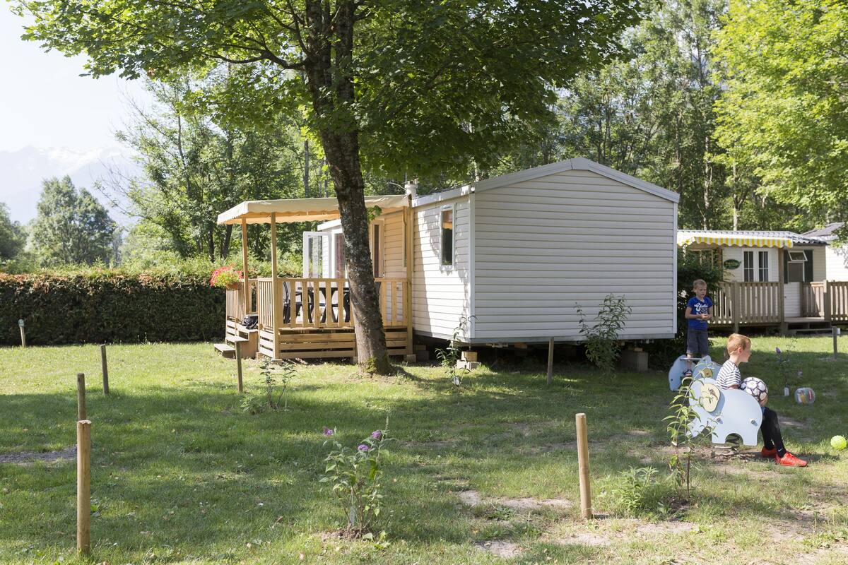 A mobile home with a porch and a small deck, surrounded by a fenced yard with grass and a few plants.