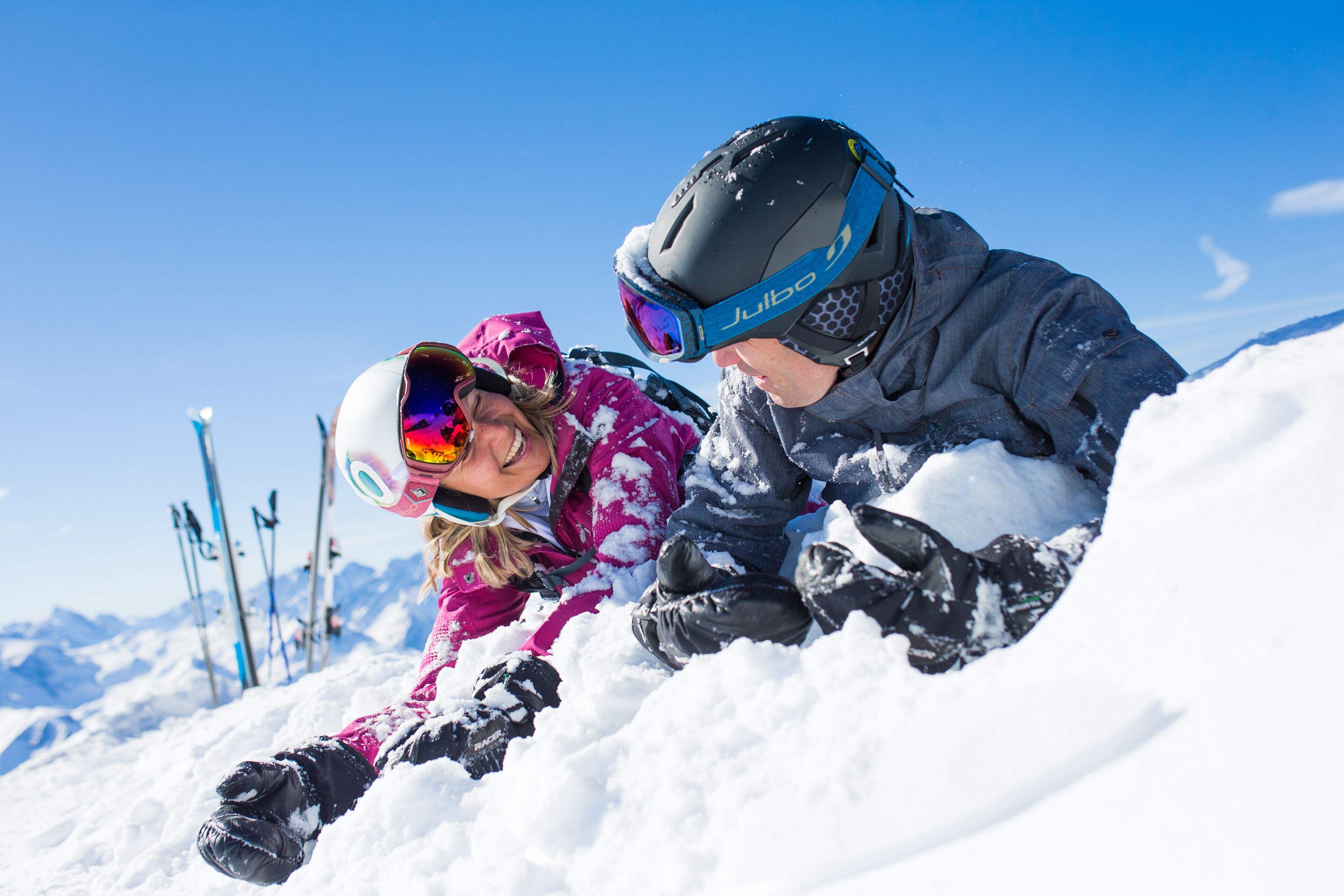 Deux personnes allongées dans la neige, avec des skis en arrière-plan.